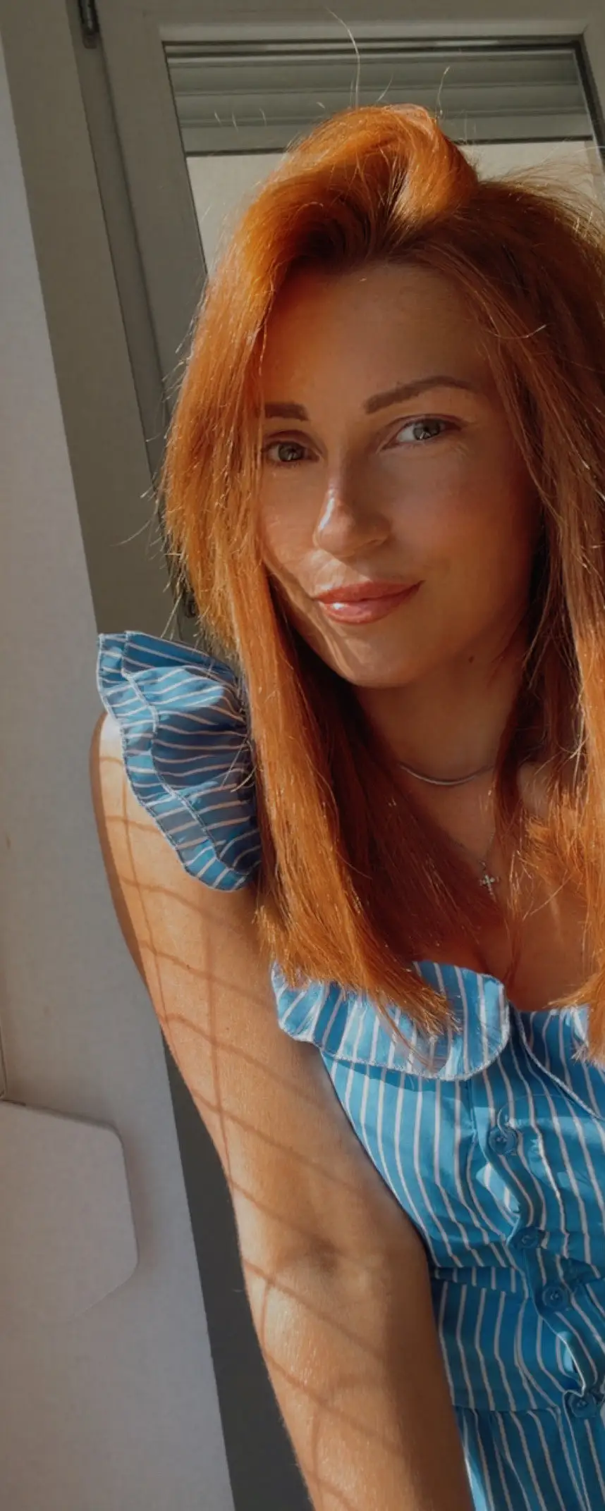 Milica beside a shuttered doorway in bright daylight, lattice shadow falling across her arm and the wall behind her.